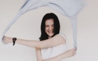 Happy woman with eyes closed joyfully waving a scarf indoors against a simple background.